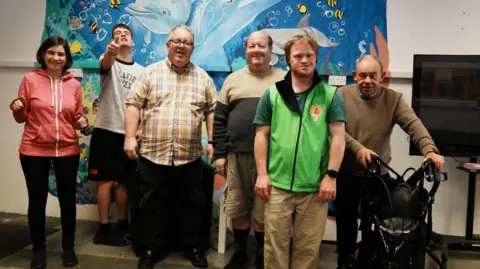 Six adults stood in front of a colourful mural on a wall. They are all smiling at the camera.