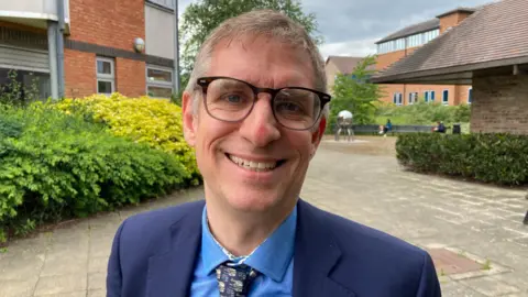 Councillor Matt Jenkins, wearing a blue suit jacket, blue shirt and patterned tie, is smiling at the camera.