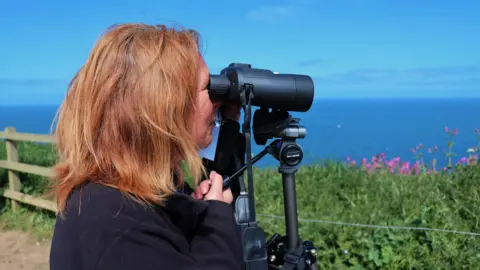 Ellen Leach A woman stands on the cliffs at Bempton, East Yorkshire and looks out to sea using a pair of black binoculars. She has long red hair and is wearing a black jacket. In the background, a wooden fence and exposed cliff is visible along with the sea in the back of the image.
