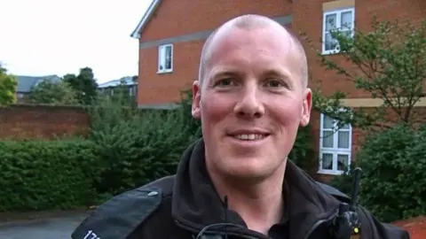 Thames Valley Police PC Jim Mahony smiling for the camera in front of a house. It is a cloudy day.