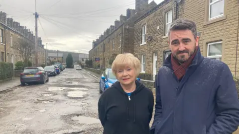 A blond-haired woman standing next to a man in a dark raincoat standing in a terraced street with lots of potholes 