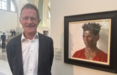 BBC/Nahem Shoa Wearing a grey a suit over an open neck white shirt, a smiling Sir Nicholas Serota is also wearing a pair of round, almost rimless glasses. He's standing alongside a portrait of the artist Nahem Shoa.