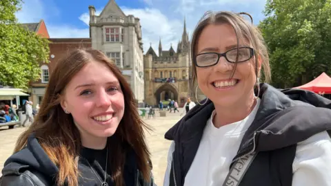 Emma Baugh/BBC Two women smiling at the camera. There are some people in the city centre in the background. The woman on the left is wearing a black leather jacket. The woman on the right has glasses and is wearing a black sleeveless jacket over a white top.
