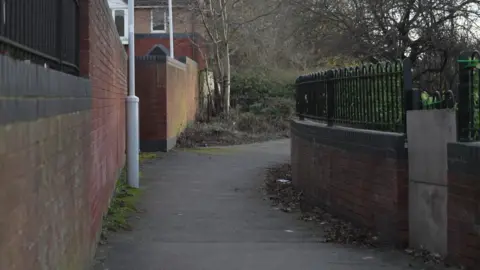 A path with brick walls on both sides, one topped with metal railings, leading to a green space lined with shrubs and trees.