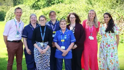 A group of eight people, some in health uniforms and some with lanyards. They are all smiling directly at the camera and they appear to be standing in a garden. There is a hedge behind them.
