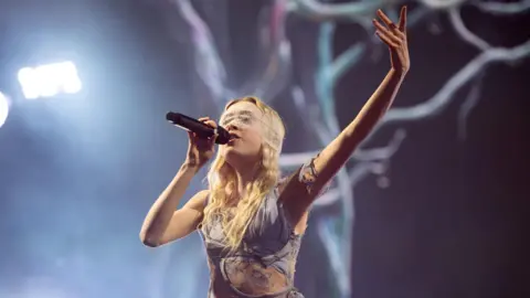 Getty Images Erin Le Count performs on stage and is holding a microphone. She is wearing a blue dress with lace cut-outs and has a lace eye-covering. 