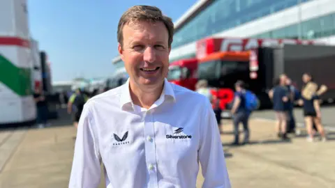 Stuart, a man with short goldish hair, wearing a white button-down shirt with a Silverstone written logo in grey on the left, smiling for camera.