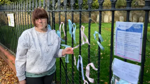 Shariqua Ahmed/BBC Adele standing next to a fence with pink blue and white ribbons and a note saying Baby Loss Awareness Week