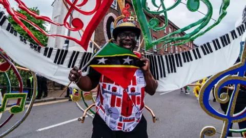 A man, wearing a colourful waistcoat, wearing a hat with musical clef on its front, with . He has large piano-themed "wings" attached to his back, with more musical notes flying above the top of the wings and to the sides. He is smiling and has his mouth open. He is part of a carnival parade. 