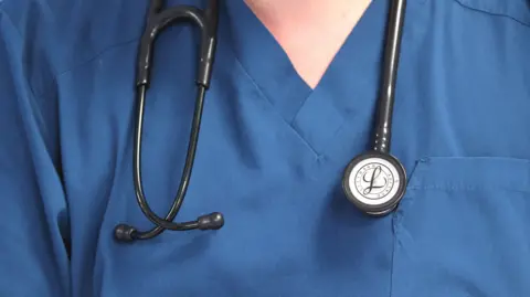 A doctor wearing a blue v-necked medical top in wearing a stethoscope around their neck, with the earpieces on one side.