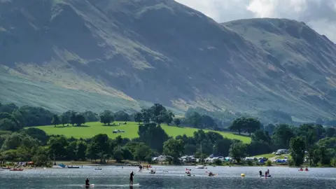 PA Media A view of Ullswater in the Lake District on a summery day. The lake is in the foreground, with people paddleboarding, kayaking and rowing. On the far bank, on a small green hill, is a campsite packed with tents and motor home. In the background, two high mountains sweep down towards the water.