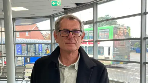 Luke Deal/BBC Cliff Waterman smiles at the camera while standing inside a bus station. He has short grey hair and is wearing black glasses with a black coat and off white shirt underneath. Behind him outside a parked double decker bus can be seen.