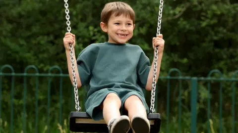 Elliot, a seven-year-old boy pictured on a swing outside. He has short brown hair and is wearing a green top and shorts. He is looking to the side and smiling. He has gaps in his teeth.