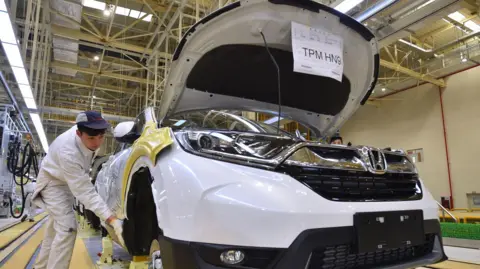 A white Honda car being made in a factory with a worker to the left of the picture wearing white overalls and a cap.