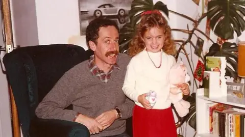 Family handout A man with short brown hair and a large moustache is smiling while sitting in an armchair in a living room. A young girl with long curly blonde hair is smiling while standing next to him. She is holding a Care Bear and a My Little Pony.