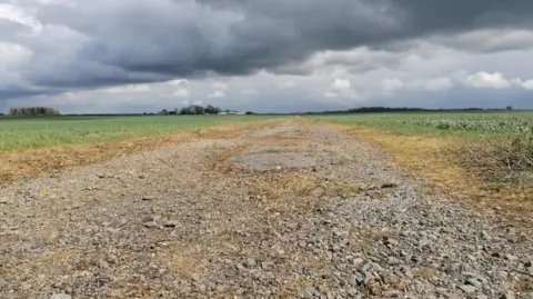 Paul Joslin/BBC The remains of the 1946 race track at Gransden Lodge Airfield in 2026. It shows a rough track, covered in broken bits of stone or concrete, with green fields on either side and grey clouds in the sky above.