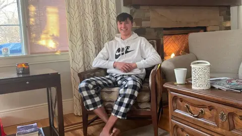 Barnaby Webber in black and white check pyjama bottoms and a sports hoodie, sitting in a chair in the corner of a room, near a lit fireplace. He is smiling at the camera and looked relaxed.