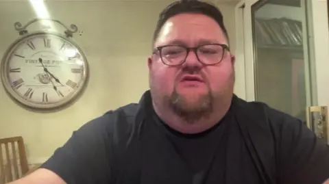 BBC James Thomas, with a clock on the wall behind him, speaking about the market. He is wearing glasses and a black T-shirt and has short, dark hair and a beard.