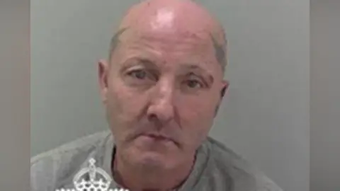 West Mercia Police Julian Thomas in a police mugshot. He has a bald head and tattoos on both sides of his head. He is wearing a grey sweatshirt.