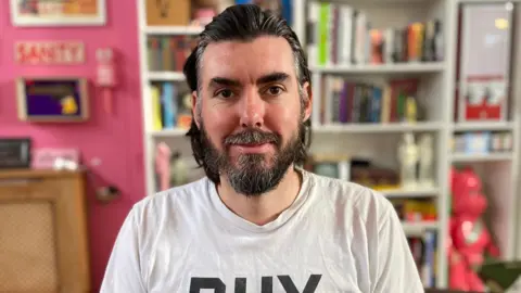 Adam Carthy has brown hair and beard. He is wearing a white T-shirt that says BHX and is sitting in front of a bookshelf.
