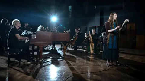 Paul Leonard-Morgan A recording session in a record studio. Paul Leonard-Morgan is sitting at the piano playing it, a group of string players are seated playing instruments and Len Pennie is reading poetry into a microphone.