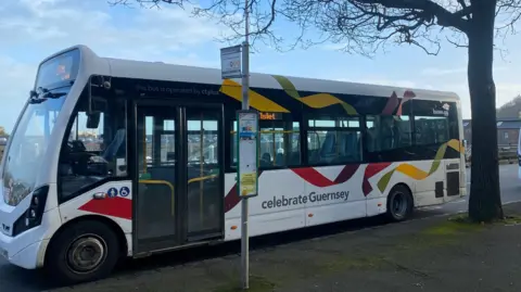BBC An empty bus is parked at a bus stop. Lettering on the bus reads 'celebrate Guernsey'. 