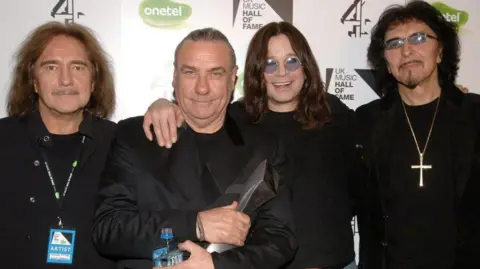 PA Media Four men dressed in black standing in front of a backdrop advertising UK Music Hall of Fame, sponsored by Onetel and Channel 4. From left to right are Black Sabbath bassist Geezer Butler, drummer Bill Ward, singer Ozzy Osbourne and guitarist Tony Iommi.