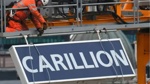 Getty Images A sign in blue and white saying "Carillion" is being lowered ground wards by a man in an orange boiler suit and white hard hat, as he is crouched on scaffolding.