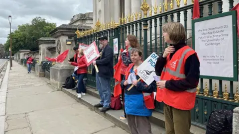 A picket line in Cambridge outside The Fitzwilliam Museum. Union members with red tabards, some holding placards, stand in a row in front of the railings outside the museum.