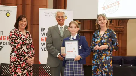 UGC A schoolgirl, in blue school uniform, holds up a certificate saying winner while at an awards ceremony. Three adults - two women and a man - are standing smiling around her. 