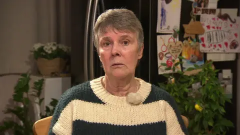 A woman with short grey hair looks at the camera with a sad look on her face. She wears a knitted stripey jumper in green and cream. Behind her is a fridge with drawings attached to it and several green plants.