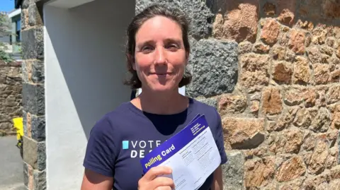 A short-haired woman in a blue T-shirt, holding polling card in the sun while she smiles.