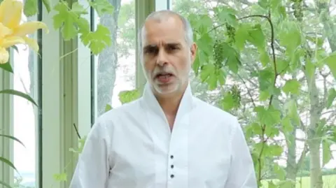 Everyone is God Party Mr White addresses the camera from inside what appears to be a conservatory surrounded by green plats. He is wearing an open necked white shirt.