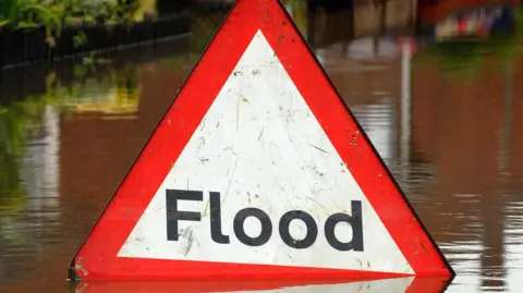 A general flood sign in flood water. It shows a triangle sign with a red outline. The work 'flood' is written in the middle of it. High flood water can be seen behind it. 