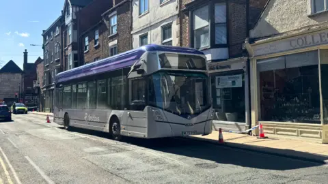A silver‑and‑purple First Bus parked on a narrow street lined with old brick and stone buildings, with traffic cones by the pavement and police tape. A police car is in the background.