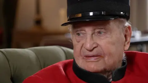An elderly man in a red uniform and black hat looks just out of frame of the camera