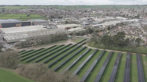 SunGift Solar Aerial view of solar panels on green field near factory and homes.