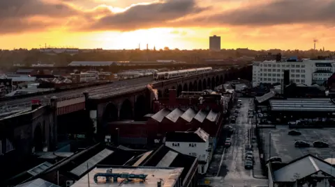 Bordesley Railway Viaduct can be seen as the sun sets in the distance. A train track runs on the left of the image with buildings and warehouses below to the right with a main street also visible.