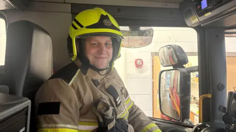 BBC Sam Heslop dressed in firefighter uniform sat in the passenger seat of a fire engine.