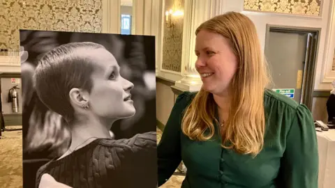 Mum Vicky Robayna, who has mid-length blonde hair, looks at a large black and white photograph of her teen daughter Liz Hatton. 