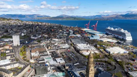 An aerial image of Greenock town centre, taken from a drone. The main high street is visible, as well as the Firth of Clyde water and hills in the distance. A large passenger cruise ship is moored in the port and two red cranes sit at the waterside.