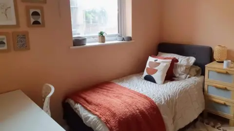 The Esther Project A peach-coloured bedroom with a single bed in a corner under a window. Next to it there is a set of drawers and in front of it there is a white desk