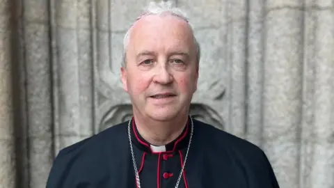 A man in black and red clerical garb stands in front of the pillars of a church. A cross is being worn around his neck. 