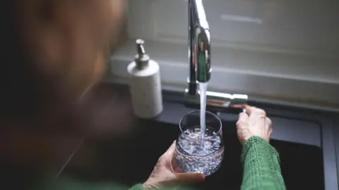 Getty Images Woman wearing a green jumper, pouring a glass of water from a kitchen tap.