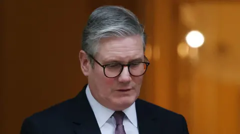 Sir Keir Starmer wearing dark suit and glasses looks down at floor