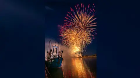 IAN WARD A fishing boat is in the foreground backlit by fireworks being launched from the bank
