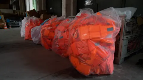 Large numbers of bright orange lifejackets are seen in plastic bags inside a warehouse