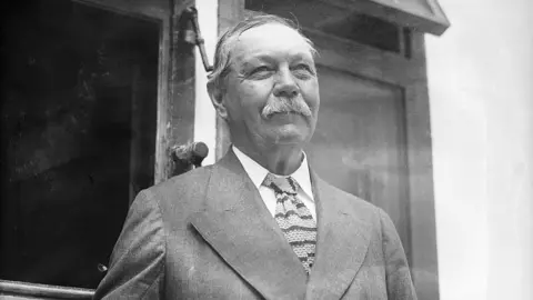 Getty Images A black and white photograph of Sir Arthur Conan Doyle dressed in a formal double-breasted suit with a patterned tie, standing in front of a building with large windows. The architectural details, such as the wooden window frame and decorative elements, suggest an early to mid-20th Century setting.