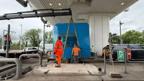 A blue toll booth being held up by a small crane and sat on a trailer. To the right of it is a black car and to the left is a crane. There is a white building above it. The sky is grey and cloudy. 