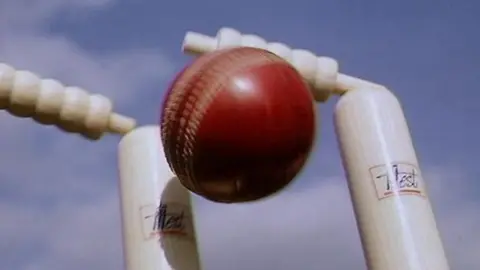 A red cricket ball hitting the white stumps, with blue sky in the background. 
